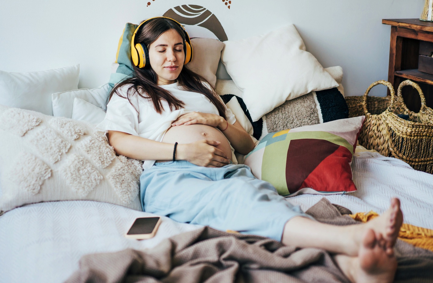pregnant person lounging in bed, wearing headphones, with their hands on the stomach.