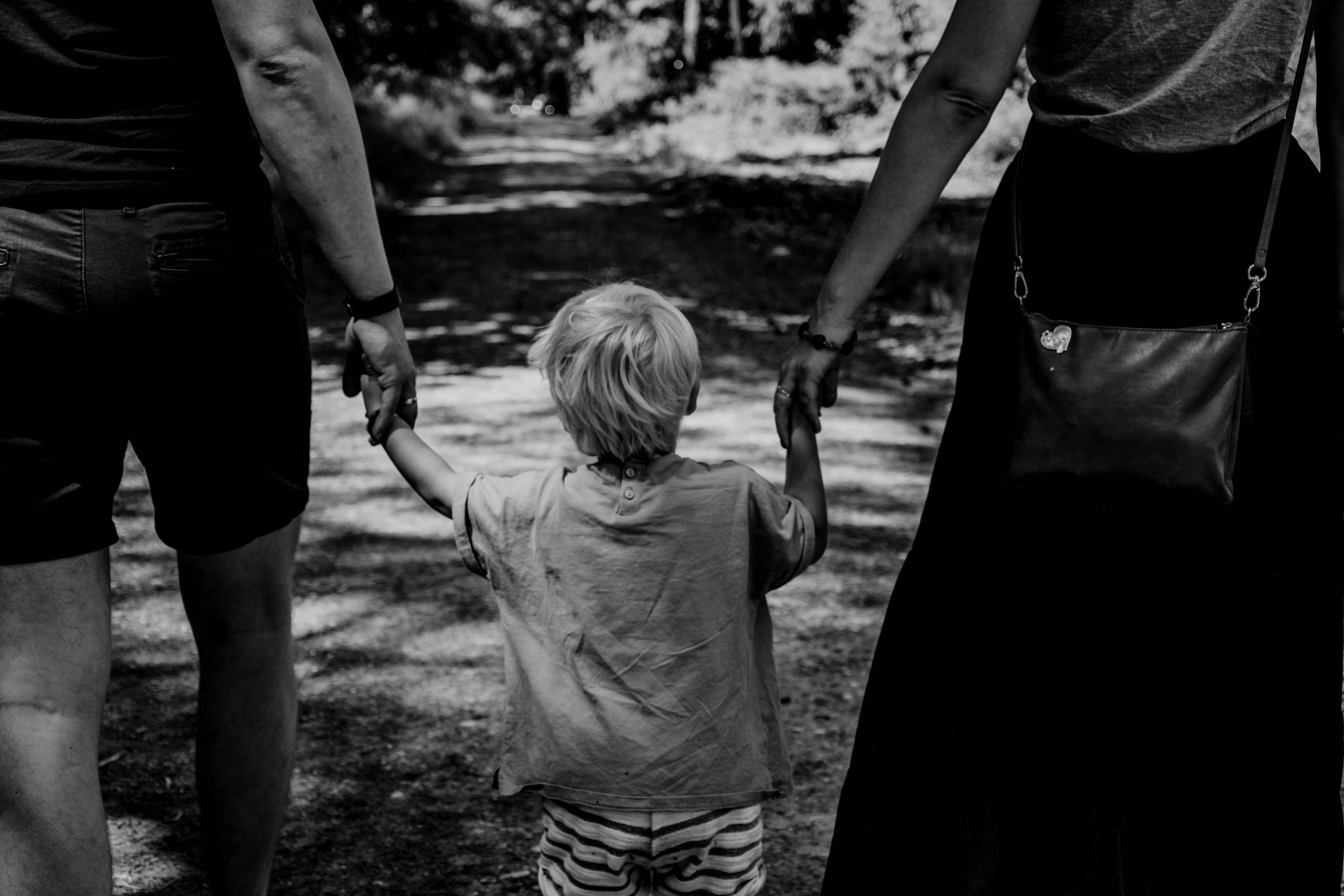 Black and white photo of a child walking holding two parents hands.
