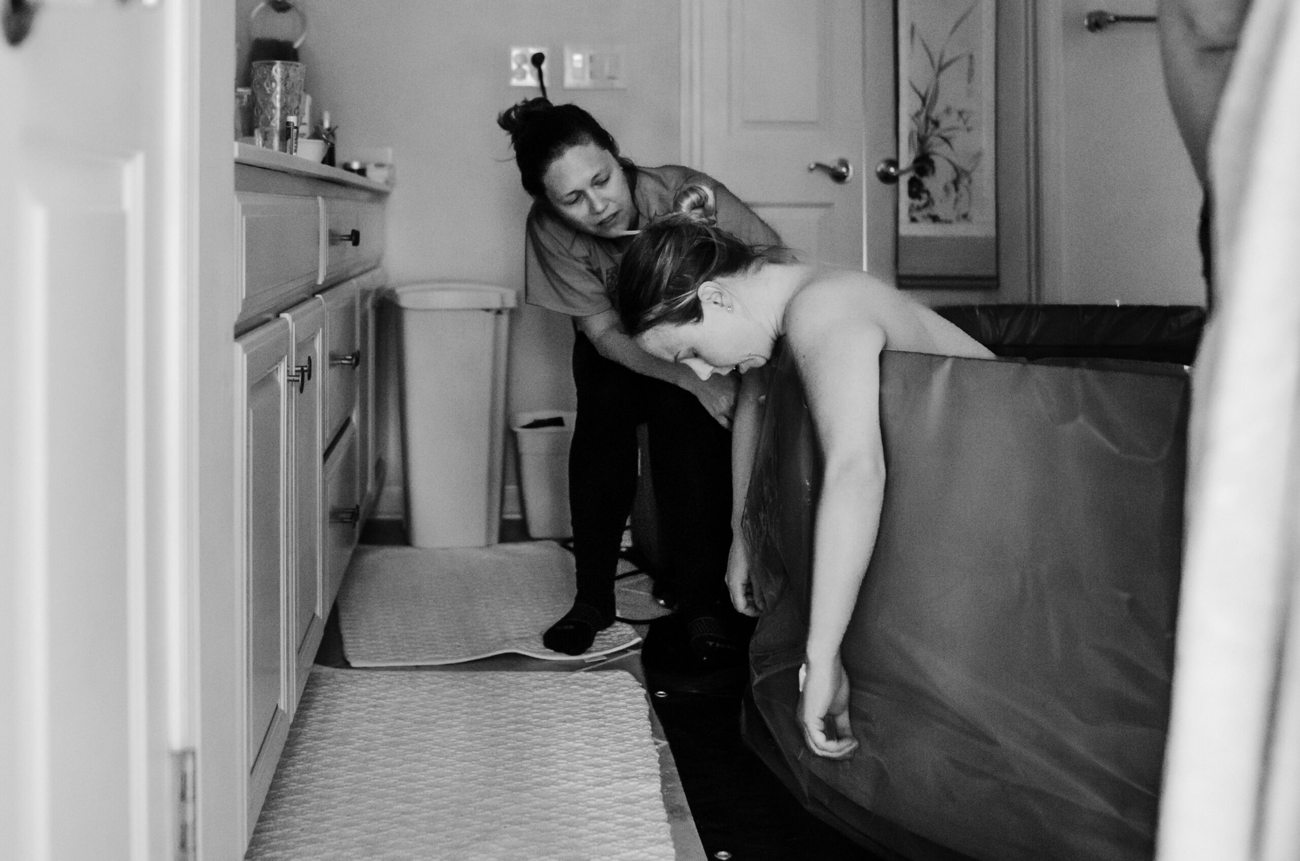 Birthing person in bath tub with doula standing next to them, offering comforting touch.