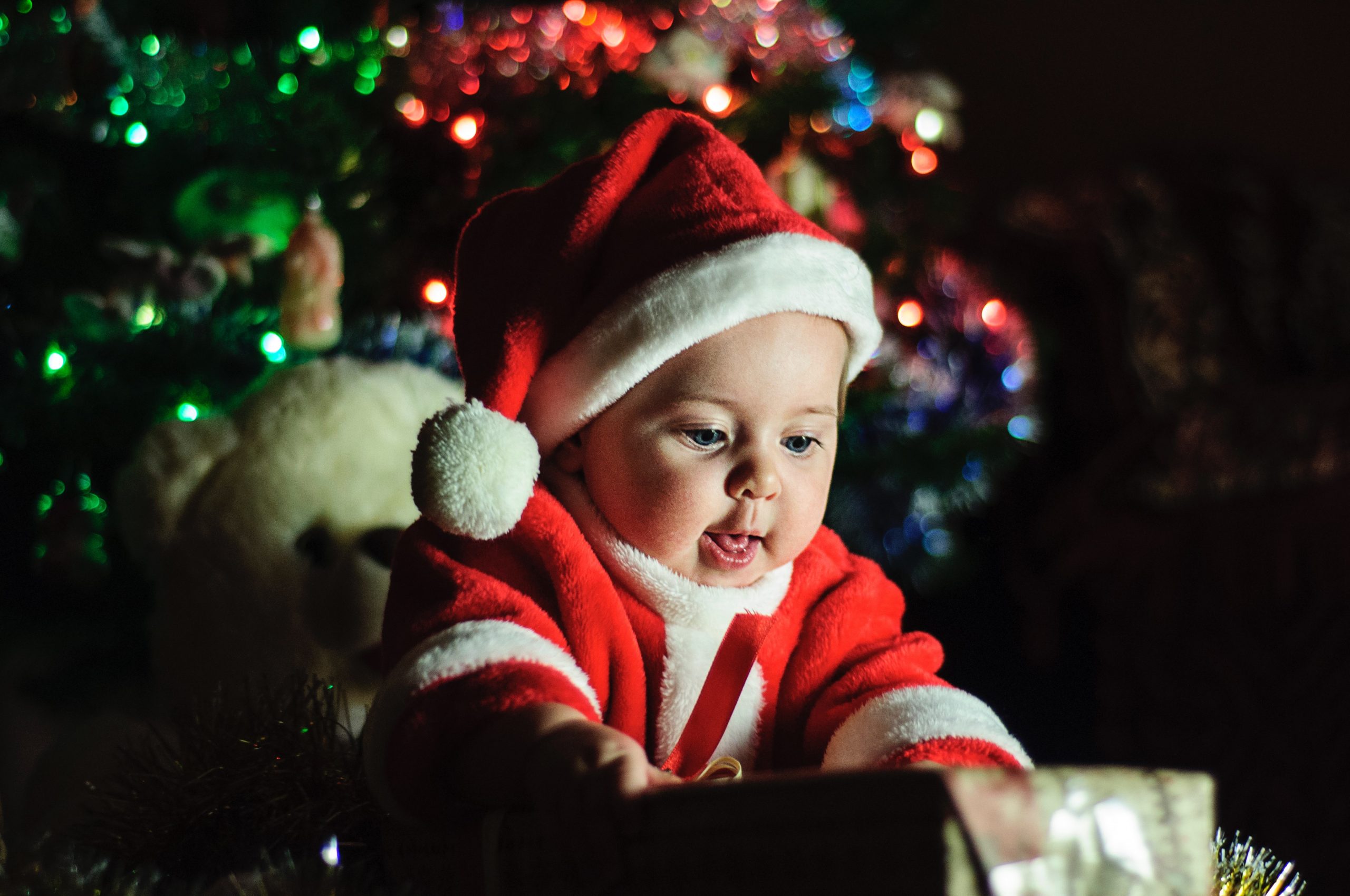 Baby wearing santa hat while opening a present in front of a holiday tree.